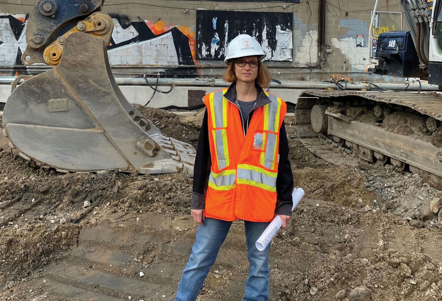 Carol Domitric wearing construction safety vest and hard hat standing in front of excavator