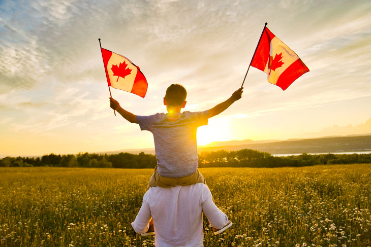 Man carrying boy on shoulders waving a small Canadian flag in each hand