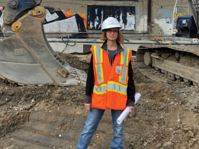 Carol Domitric wearing construction safety vest and hard hat standing in front of excavator