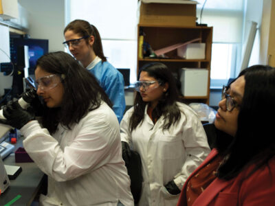 Siddiqua conducts research in lab with three others in white lab coats. One of them is looking in a microscope