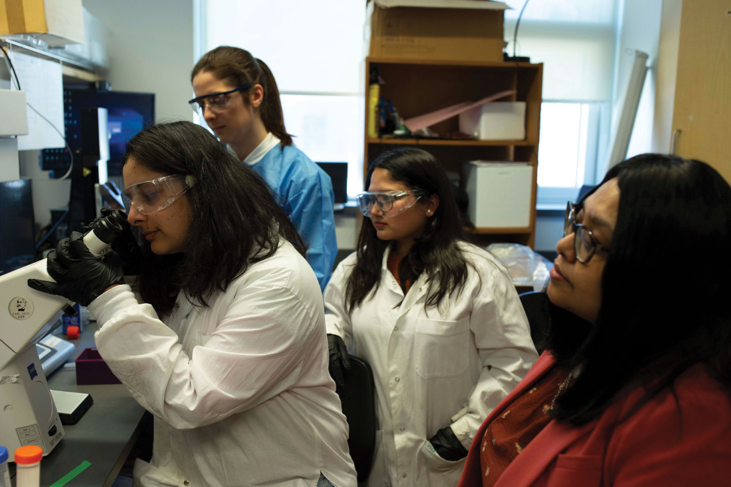 Siddiqua conducts research in lab with three others in white lab coats. One of them is looking in a microscope