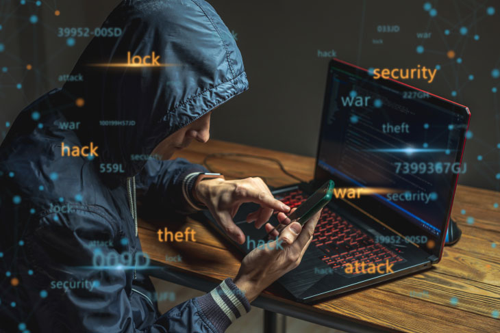 A hacker in a hood with a phone is typing on a laptop keyboard in a dark room