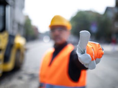 Worker wearing a safety vest and hard hat raises hand in a stop or directing gesture at a construction site. Heavy machinery is visible in the background, indicating ongoing work