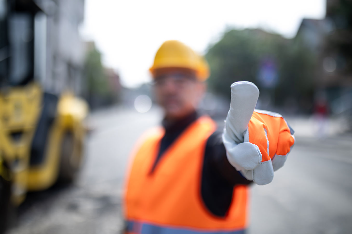 Worker wearing a safety vest and hard hat raises hand in a stop or directing gesture at a construction site. Heavy machinery is visible in the background, indicating ongoing work