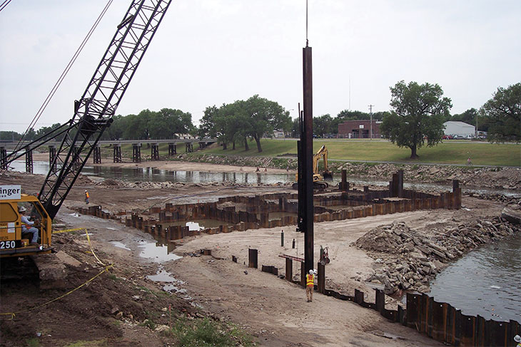 Crews drive steel sheet piling to form a cofferdam in riverbed